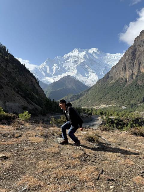       Hiker crouches playfully on mountain viewpoint surrounded by pine forests and snowy peaks.
  