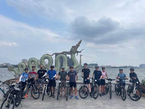       Cycling group lines up with bikes in front of dragon sculpture beside large lake in Hanoi.
  