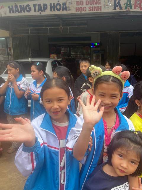       Cheerful Vietnamese schoolchildren wave enthusiastically at the camera outside a shop.
  