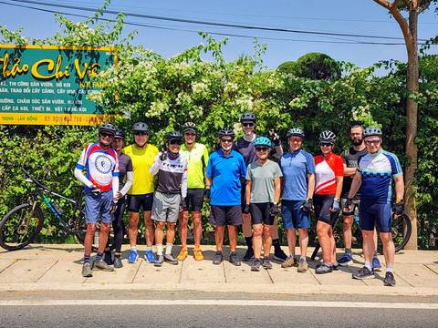       Large group of cyclists posing together in front of greenery and a Vietnamese welcome sign.
  