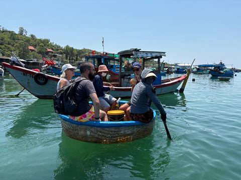       Travelers sit in a traditional round basket boat on turquoise water surrounded by colorful fishing boats.
  
