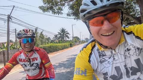       Selfie of two smiling cyclists riding down a rural Vietnamese road lined with palm trees.
  