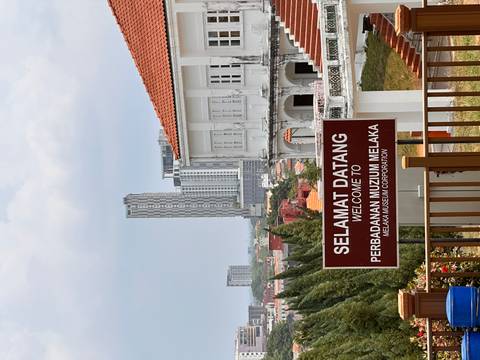       Sign welcoming visitors to the Melaka Museum Corporation with city skyline behind.
  