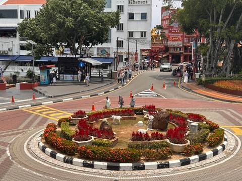       Elevated view of a roundabout with red flower beds and tourists exploring Melaka streets.
  