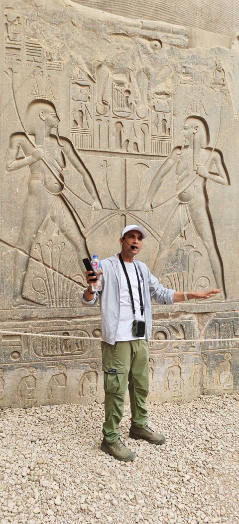       Egyptian guide speaking through headset microphone in front of carved hieroglyphic stone reliefs.
  