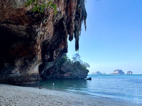       Sheer limestone overhang framing a quiet cove with shallow emerald water; a couple of swimmers explore the beach.
  