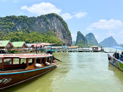       Floating village and long-tail boats moored against towering limestone cliffs in a green bay.
  