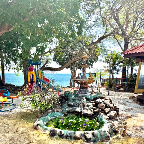       Small seaside playground with colorful slides and a decorative fountain under leafy trees overlooking the ocean.
  