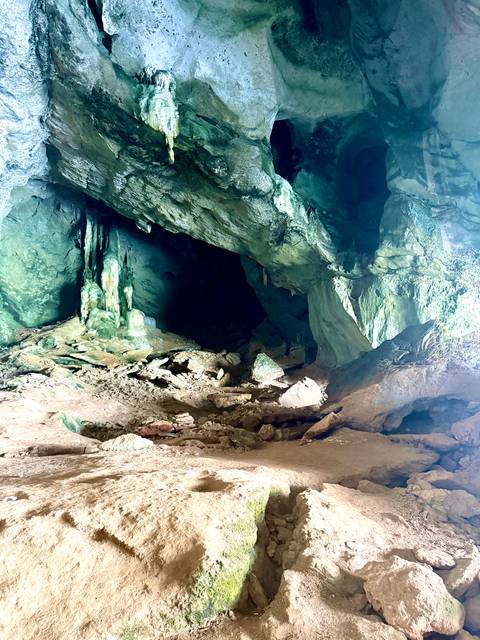       Dimly lit limestone cave interior with stalactites, green mineral stains and rocky floor.
  