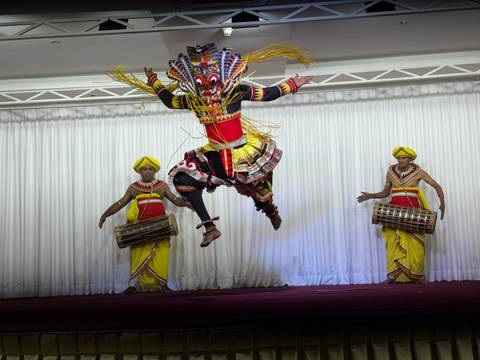       Energetic traditional Kandyan dancer mid-air with vibrant costumes accompanied by two drummers on stage.
  