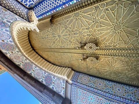       Ornate golden door with geometric patterns and vibrant zellij tilework framing the entrance to a Moroccan palace.
  