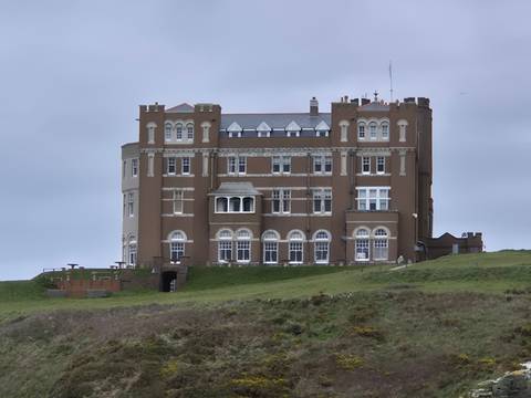       Large brown stone building resembling a coastal castle sits on a grassy hill under overcast skies.
  