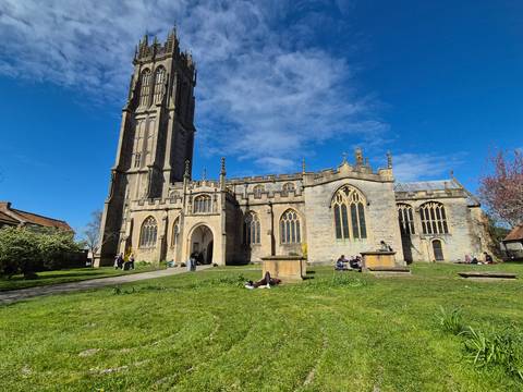       Stone church with tall tower, arched windows and people lounging on the grassy churchyard under blue skies.
  