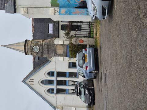       Tall narrow clock-tower building with parked cars in a small village setting under grey clouds.
  