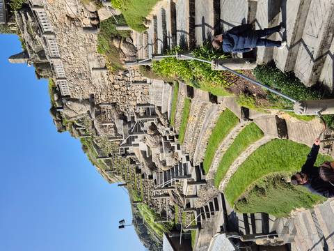       Stone terraces and steps of the open-air Minack Theatre carved into a cliffside with visitors exploring.
  