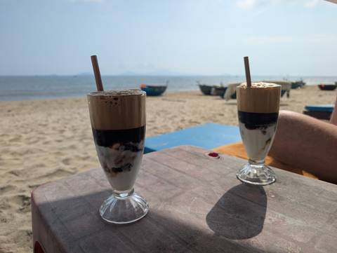       Two tall iced coffees set on a rustic beach table overlooking a calm sandy shoreline
  