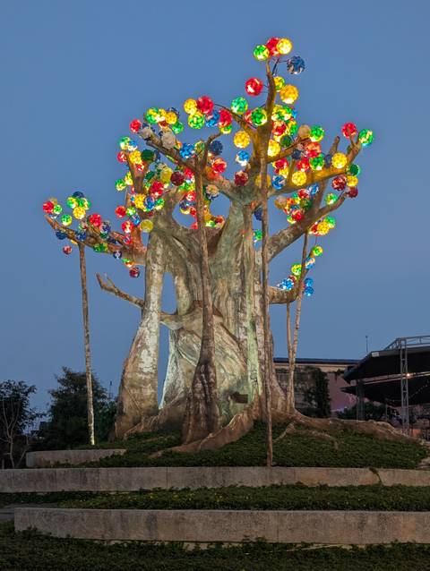       Public art sculpture of a tree with multicolored illuminated flowers against evening sky
  