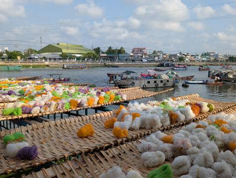       Bundles of colourful rice noodles drying on woven racks beside a bustling Mekong river port
  