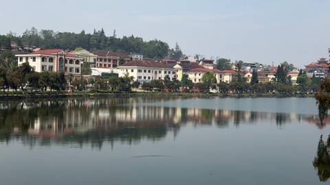       Calm lake reflecting colonial-style buildings and green hills in a Vietnamese highland town.
  