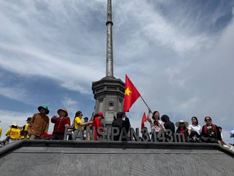       Crowd of visitors waving Vietnamese flags at the Fansipan summit marker under blue skies.
  