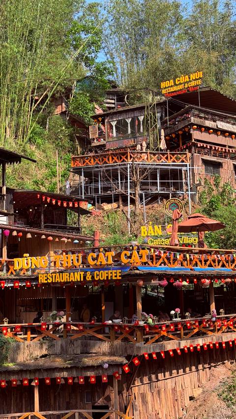       Colorful signage and hillside wooden structures marking entrance to Cat Cat Village.
  