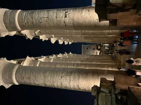       Night-lit colonnade of Luxor Temple with visitors walking below towering pillars
  
