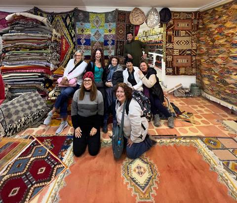       Group of women pose inside a colorful carpet shop surrounded by stacked rugs
  