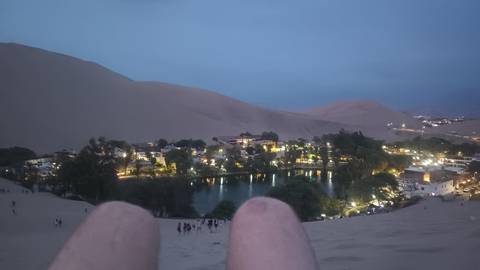       Evening view over a desert oasis village with lit palm-lined lagoon, seen from a sand dune with traveller’s knees in foreground.
  
