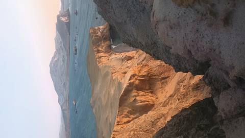       Vertical coastal cliff of ochre rock jutting into turquoise sea with distant islands under pastel sky.
  