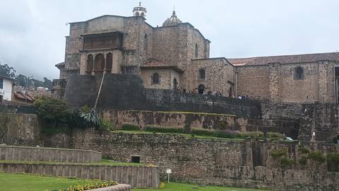       Stone Inca-Spanish church complex with dark retaining walls and bell tower under cloudy sky.
  