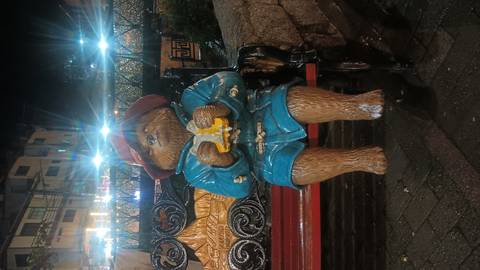       Nighttime photo of a Paddington Bear statue sitting on a bench eating fruit under bright streetlights.
  