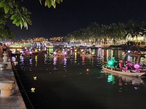       Night scene of brightly-lit boats carrying visitors with colorful lanterns along a palm-lined river.
  