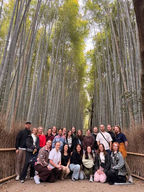       Large tour group posing beneath the soaring stems of a tranquil bamboo forest.
  