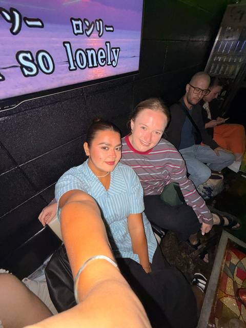       Casual overhead selfie of two friends seated indoors with others blurred behind them.
  