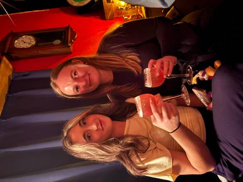       Two friends toast with cocktails in a cozy, warmly lit bar.
  