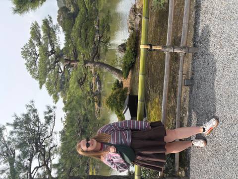       Traveler posing beside a manicured pond and sculpted pine trees in a Japanese garden.
  