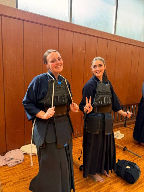       Two smiling women wearing kendo armor posing inside a wooden dojo.
  