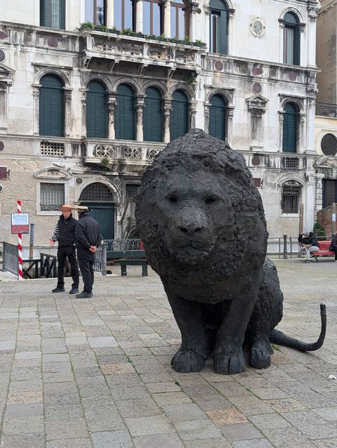       Textured black lion sculpture in a Venetian square with two gondoliers chatting nearby.
  