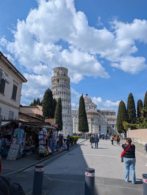       The Leaning Tower of Pisa and cathedral framed by tall cypress trees under a vivid blue sky.
  