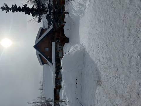       Snow-covered log lodge under a bright hazy sun with thick drifts surrounding the entrance.
  