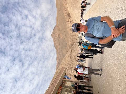       Tourists gather on a dusty plateau backed by barren limestone hills; one man in sunglasses poses in the foreground.
  