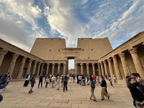       Wide courtyard of an ancient Egyptian temple lined by tall columns and bas-reliefs under a partly cloudy sky, filled with visitors.
  