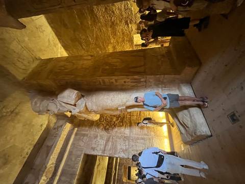       Traveller poses beside a colossal pharaonic statue inside a dimly lit rock-cut chamber with carved walls.
  