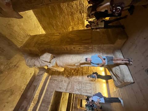       Similar interior temple scene with tourists photographing a giant carved statue under yellow lighting.
  