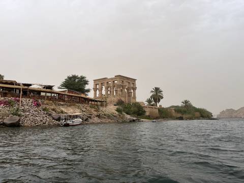       Ancient stone temple of Philae rises beside the Nile, viewed from a boat on choppy water under hazy skies.
  