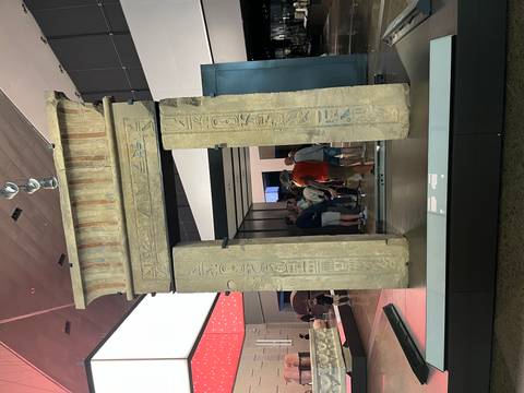       Stone gateway with hieroglyphs inside a museum hall where a small tour group listens to a guide.
  