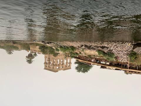      View from the water of the ancient Kiosk of Trajan on a rocky riverbank with palms.
  