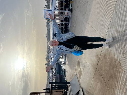       A smiling woman raises her arms on a pier with boats and sunrise light in the background.
  