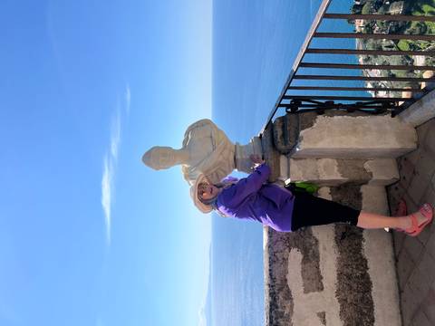       Traveler hugging a marble bust on a high balcony with expansive sea views.
  