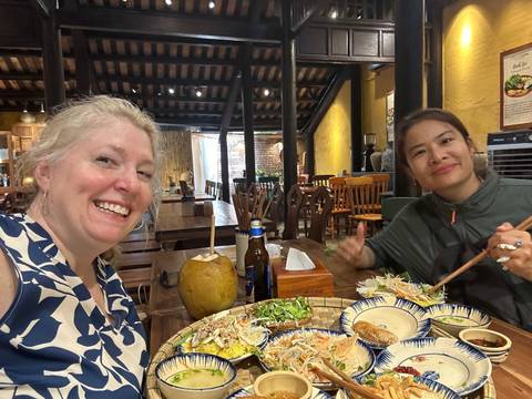       Two women enjoying a Vietnamese feast at a wooden restaurant table, smiling at the camera.
  